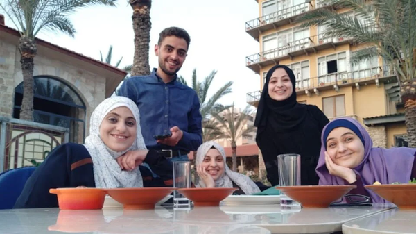 An image of a smiling young man and four smiling women, all facing the camera. There is a table with food bowls in front of them. In the background are tall date palms and multi-story buildings. 