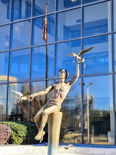 In front of a glass window building, and next to an American flag, a happy statue of a shirtless young man playing at a pier, seated atop a piling, where several gulls frolic and land in his hands.