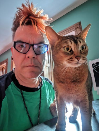 Middle-aged white man with spiky orange hair and green T-shirt looks into camera next to large ruddy Abyssinian cat