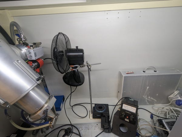Photograph of the inside of an x-ray cabinet. There's a fan and a result stand pointed at the turbo pump on a large silver x-ray gun on the left side of the image towards the right of the image is the turbo pump, vacuum gauge and a collection of wires
