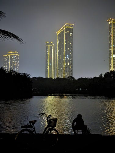 A late night photo on the outskirts of exurban Hanoi. We're standing behind a man who squats on the banks of a lake where he fishes alone. We see he and his parked bicycle in silhouette. Their images stand out sharply against the shimmering light of apartment blocks across the lake. We can make out for such towers each appearing to be thirty or more floors.