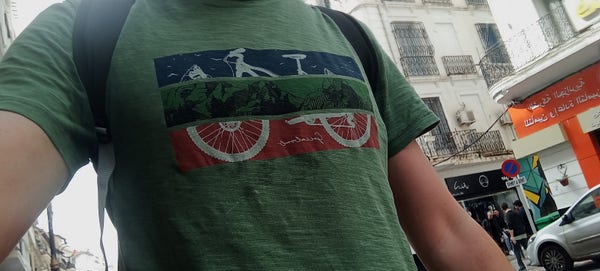 Mézigue portant un t-shirt vert avec vélo (un VTT) sur paysage de montagne. Le décor en arrière plan est celui d'une rue d'Alger : des immeubles, des petits commerces, des bagnoles ! 
