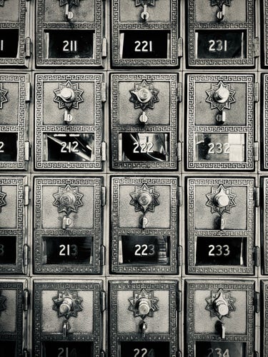 A black and white photo of a patterned array of numbered post office boxes with ornate details and dials. They are still in active use.
