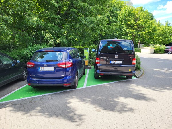 Zwei Autos stehen auf grün markierten Parkplätzen, zwischen den beiden Fahrzeugen ist eine Ladesäule zu sehen. Beide Autos sind nicht an die Ladestation angeschlossen und sind auch keine E-Autos.