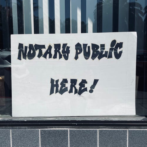 White sign in a window that says "Notary public here" in a black graffiti font.
