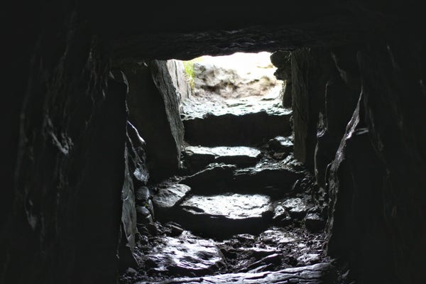 Inside an Irish burial tomb. The image is dark at the top and sides. Light comes through a square entrance built of rock, with shallow, weathered steps leading down to the bottom center of the photo. There is a slight sheen of moisture from the cold, damp air.  The only light source is outside.