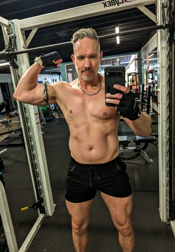 Gym mirror selfie wearing short, tight black shorts showing off the quads, and shirtless, holding up my left arm and flexing the bicep