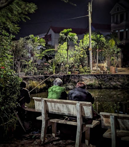 An evening photo of three men sitting around a village pond while one of them fishes. The pond, in Hưng Yên Province, not far from Hanoi, has stone and concrete reinforced banks, and is, in fact, part of a series of ponds that ring this village. The village was once accessible only by a series of small bridges. Two of the men sit on a concrete bench facing away from us while the third is crouching on the bank with a pole in hand. Across the water we can see one of the old bridges covered in potted trees and benches. In the blurry, more distant background, we can see the tops of some village homes, large and modern looking.