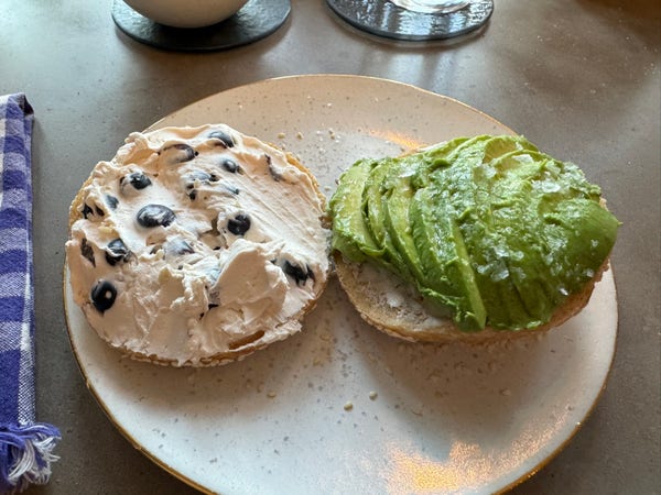 A sesame seed bagel cut in half. One half is topped with cream cheese with fresh blueberries mixed in. The other half is topped with half an avocado with flaky sea salt. 