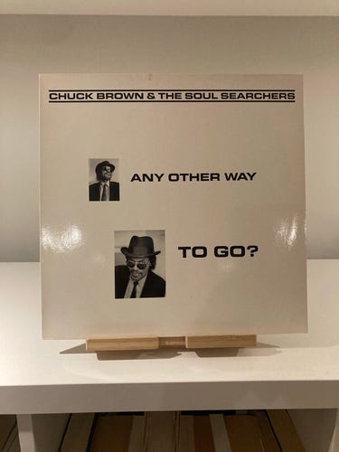 Vinyl record sleeve for "Chuck Brown & the Soul Searchers - Any Other Way to Go?", with black text on a white background, and two black and white photos of the artist, displayed on a wooden stand.