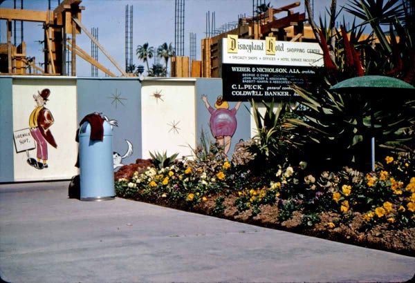Construction site at Disneyland Hotel with a sign, cartoon figures painted on a wall, a trash can, and flowers in the foreground. 