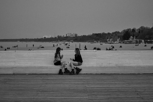 Two girls sitting in the middle of a long bench on the pier. Turned in their direction and talking animatedly.