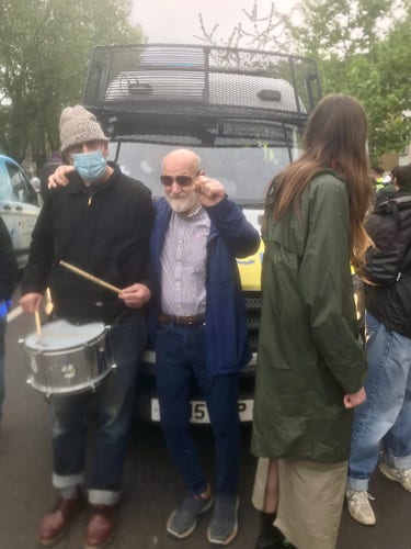 Standing in front of a Met Police van trying to transport refugees to a prison barge is Dave, in jeans and sunglasses, standing next to a man with a drum and some other protesters, who successfully prevented the Home Office during their brutal crackdown on humans trying to find safety.