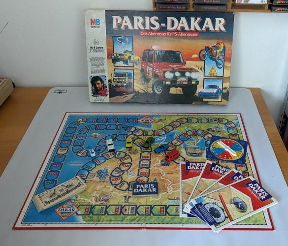 Aufbau des Brettspiels Paris-Dakar. Im Hintergrund der Karton, im Vordergrund das Spieldfeld mit Karten, Spielgeld, Autos und einem kleinen Hubschrauber.