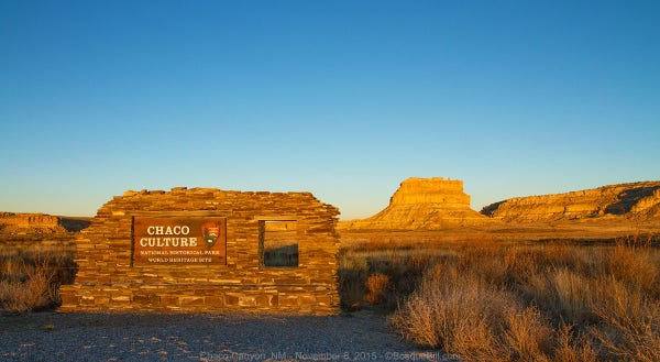 Golden afternoon light illuminates the entrance sign to Chaco Canyon and a butte behind. The sign is built of stacked rock to simulate the ancient ruins behind.
©BosqueBill.com