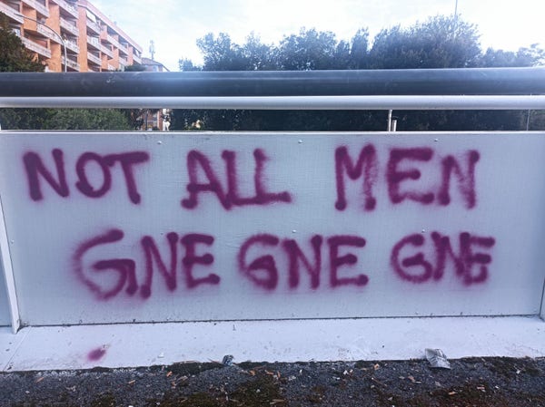 Scritta fucsia su un muretto di cemento bianco "NOT ALL MEN GNE GNE GNE"