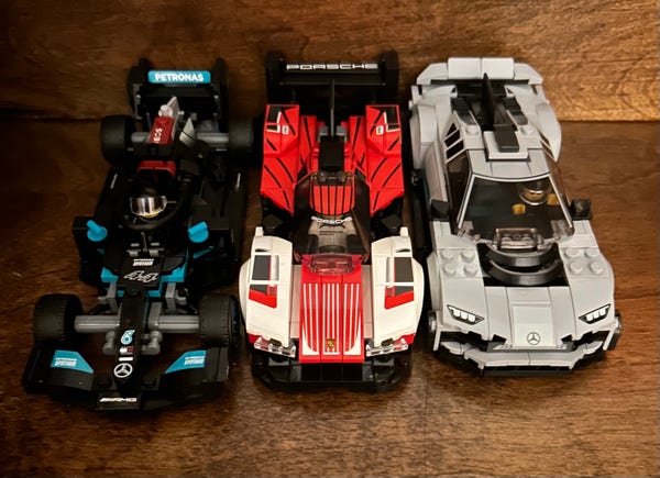 Three toy race cars representing a Mercedes W12 Formula 1 car, a Porsche 963, and a Mercedes-AMG Project One, arranged side by side on a wooden surface.