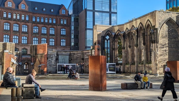 Foto der Gedenkstätte St. Nikolai in der Hamburger Innenstadt. Menschen sitzen auf Bänken, Informieren sich auf Schildern, es ist ein wolkenfreier Tag im Sommer.