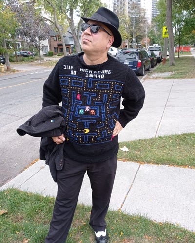 A man in a fedora and sunglasses wearing a hand knit sweater with a Pac-Man video game pattern on it, complete with a high score of 16440.