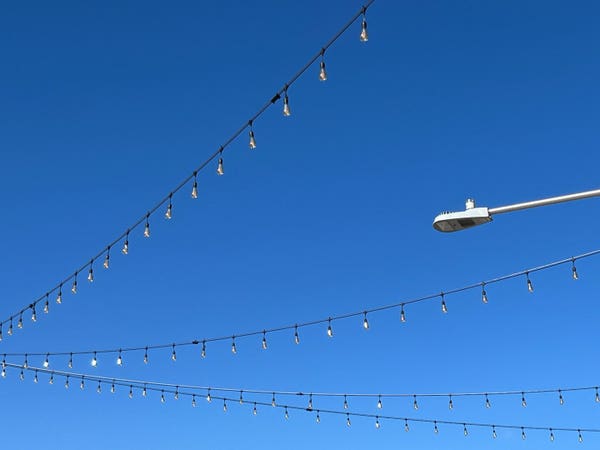Three strings of large lights over a street against a blue sky, with a street light on the side.