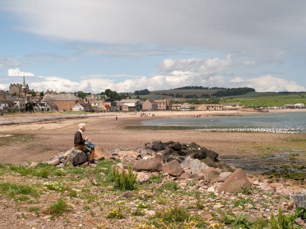 An old woman sitting on a stone by a beach. A village in the background.