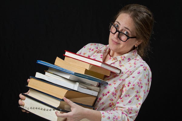 Me wearing black plastic framed glasses and a nose ring, leans over while carrying a stack of books against a black background. I am a white woman with my hair in a braid and a white floral print shirt.