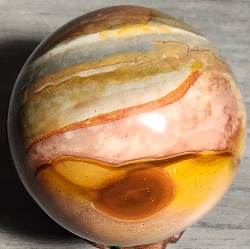 It's a sphere of Polychrome Jasper with horizontal stripes and swirls similar to the bands on Jupiter the planet. In the lower center of the sphere is a reddish-brown oval. There are reds, yellows, pinks, grays, and blues throughout the sphere in bands and swirls. The background is muted gray. There is a tiny bit of the stand showing at the bottom of the picture.
