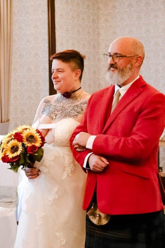 Me walking Taylor down the aisle. She’s holding a bouquet of roses and sunflowers, and is wearing a white dress with a slashed front revealing her upper chest is tattooed. She has close-cropped auburn hair and is smiling, looking out of frame at her wife. I’m wearing a dark kilt and red jacket, and I have a mostly-white beard and a bald head. I’m smiling too!