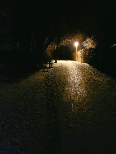 Photograph of a walking path surrounded by trees at night. The ground is covered with ice, while a yellow lamp down the path provides the only visible light source. 