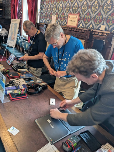 People working together to fix broken electronics.  There's various bits of kit on a long table, and it focuses on two people working together fixing a laptop.
