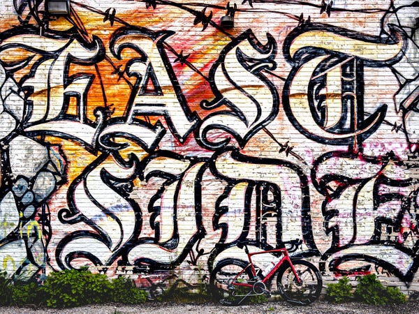 Photo of a red BMC roadmachine road bike standing next to a brick wall that is covered in a large graffiti style mural. Mirror features the words “east side” in large stylized black letters against a white background. there are some strings of barbed wire behind the words. 