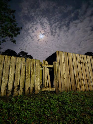 View up a small hill where an old, weathered wood-slat fence, in need of repair, stands atop the green grassy hill, beneath a cloudy blue-grey sky illuminated by the moon's glow behind the cloud cover.
