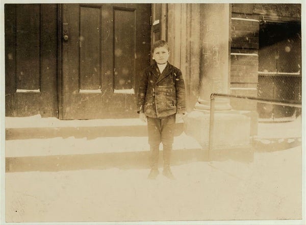  The image depicts a young boy standing in front of a doorway. He is dressed in a dark-colored outfit, possibly a suit or a jacket with trousers, and has his hands by his sides. His expression is neutral, and he appears to be looking directly at the camera. The background suggests an urban setting with what looks like steps leading up to a building, indicating that the photo might have been taken in a city or town. There is no visible text on the image, and it has a sepia-tone effect, giving it a vintage or historical feel.
