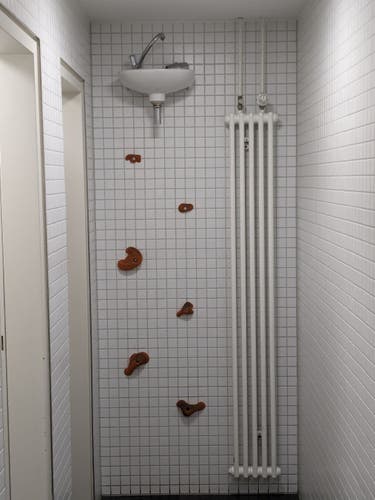 Weiß gekachelte Wand in einem Vorraum einer #Toilette.An der der Wand hängt ganz oben, knapp unter der Decke, ein Waschbecken. Um das Waschbecken erreichen zu können, sind hilfreicherweise rote Klettergriffe, wie an einer #Boulder-Wand angebracht.
#PublicToilet
#ToilettesDuMonde