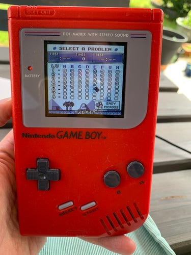A red Nintendo Game Boy held in a hand with the screen displaying the game menu for "Select a Problem" in a puzzle game.