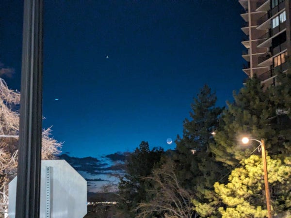 Photo taken a night of a very slim crescent moon. The back side of a stop sign is prominent in the foreground, as though the photographer has no idea how to frame a photo.