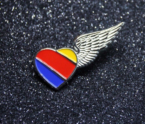 Enamel pin of a heart with colored stripes (yellow, red, and blue) and a wing attached, on a glittery black background.