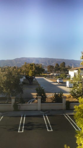 Photo of a parking lot with a masonry-and-steel fence at the perimeter. Behind the fence a concrete street with a few trees and buildings is visible. In the distance a mountain ridge is silhouetted against the clear blue sky.