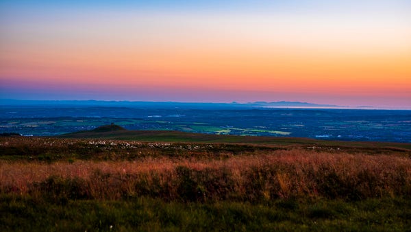 A evening dusk with the sun setting and clear sky over looking the North West of Liverpool and North Wales next to the sea. The ground is green. The sky is a mix of blue red and yellows 