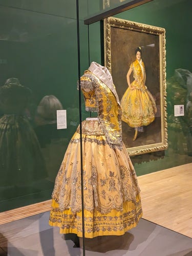 Colour photograph of a yellow dress with a very full, long skirt, displayed in a glass case. On the wall behind is a portrait of a woman wearing the dress. 