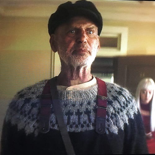 Scene from Dead Boy Detectives.
Picture of the lighthouse keeper in the office. He's an older man with grizzled short beard, black cap, and a glare in his eyes. He's wearing a black and white sweater. The yoke is white or gray with black crosses circling the yoke. 
In the background is Niko.