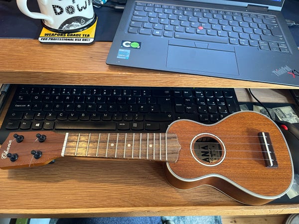 Photograph of a small wooden ukulele lying on a desk in front of a computer keyboard. A grey laptop PC and a mug of tea are visible on the shelf above.