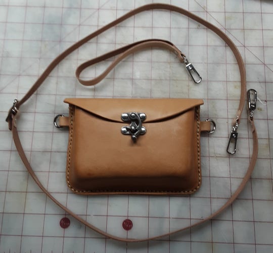 A undyed, vegetable tanned leather bag that converts from belt pouch to clutch to cross body bag. The front is a molded rectangular pouch and the back is flat. It has a belt loop on the back and "D" rings on either side for the optional wrist strap or cross body strap. It closes with a flap secured with a decorative gun metal grey  (dark silver) swing clasp. 