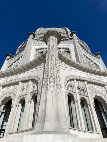 Exterior of an ornate building with intricate architectural details and arched windows against a clear blue sky. The corner of the building at the center of the photograph features the symbols from the world's major religions.