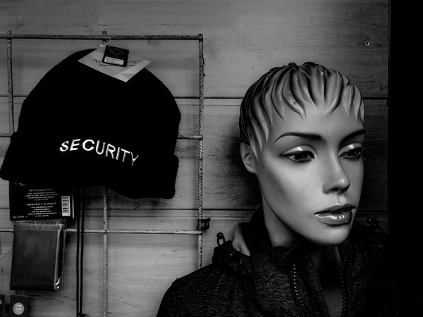 Black and white photo of a detial of a shop window featuring a black toque on the left with the word "SECURITY" stitched in white letters and a female mannequin head with short hair on the right.