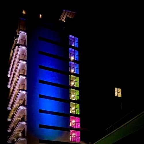 Nighttime shot of a hotel with different colored lights on the floors (3 purple, 3 green, 3 pink) along with a single window illuminated in yellow off to the side.