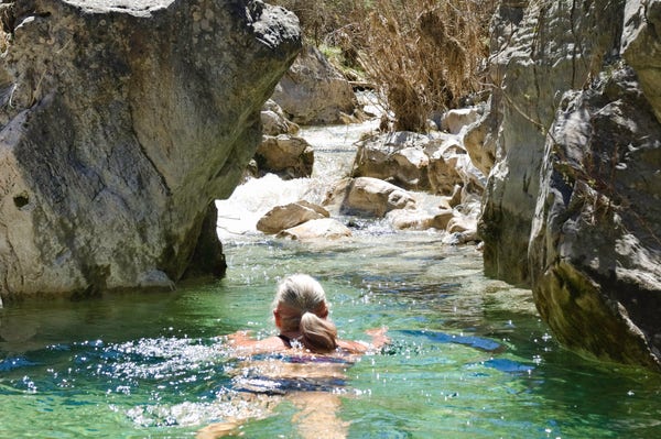 A person swims in a turquoise rock pool with steep rock on either side. Beyond is a small waterfall