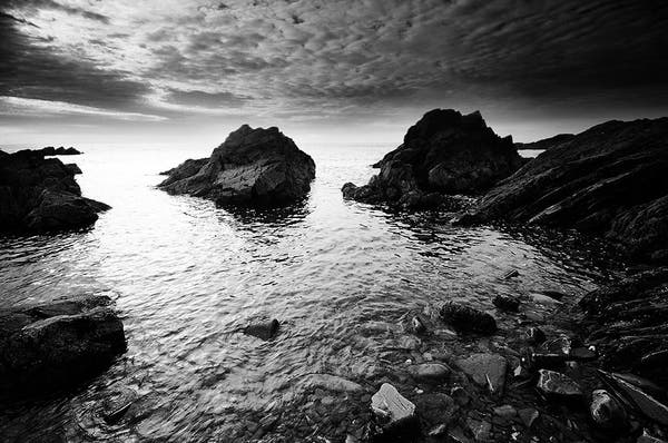 Atmospheric black and white photo of a small rocky cove, with a rocky foreshore and clouds over head.