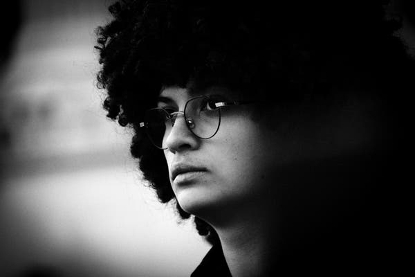 A black and white close up photo of the face of a young man with a full head of frizzy hair and wearing round glasses in the style that is sometimes known as John Lennon glasses. Hi hair is thick and black and merges into the whole of one side of the photo.