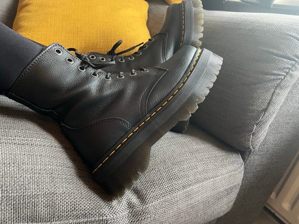 A pair of black Doc Martens boots with yellow stitching and platform soles, on a gray couch with a mustard yellow cushion in the background.
